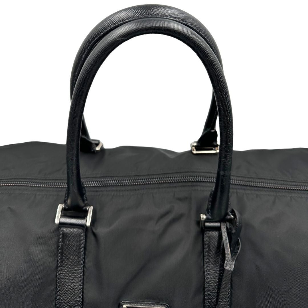 Manici di un borsone Prada in nylon nero con finiture in pelle Saffiano tono su tono e parti metalliche argentate. Completo di dustbag, tag porta address e lucchetto. Indossabile a mano.