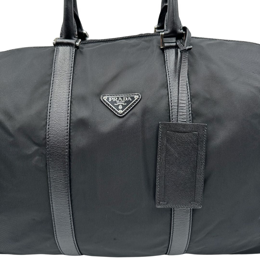 Front di un borsone Prada in nylon nero con finiture in pelle Saffiano tono su tono e parti metalliche argentate. Completo di dustbag, tag porta address e lucchetto. Indossabile a mano.