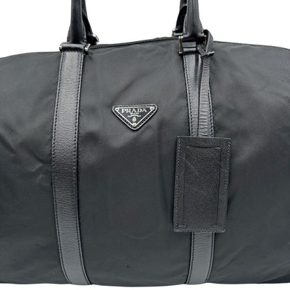 Front di un borsone Prada in nylon nero con finiture in pelle Saffiano tono su tono e parti metalliche argentate. Completo di dustbag, tag porta address e lucchetto. Indossabile a mano.