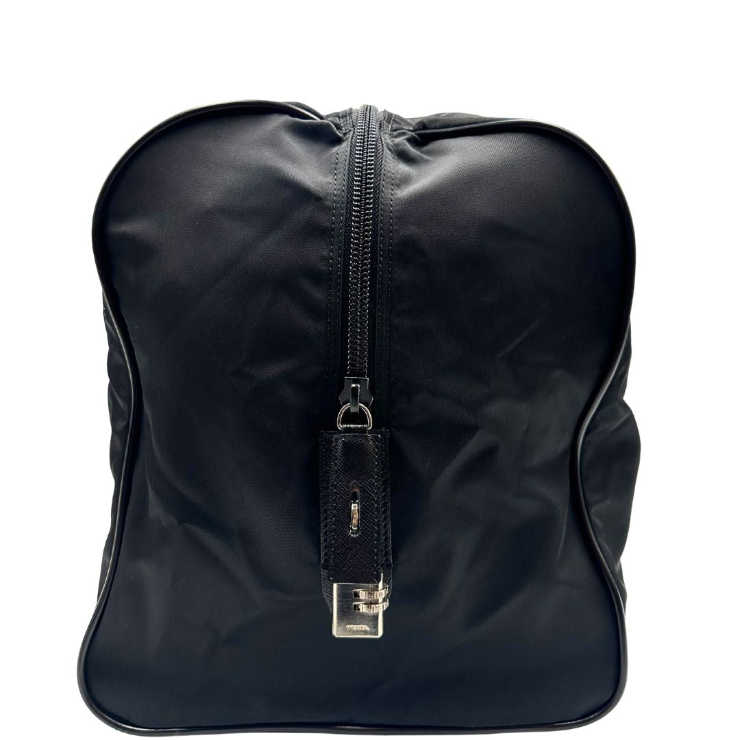 Vista laterale di un borsone Prada in nylon nero con finiture in pelle Saffiano tono su tono e parti metalliche argentate. Completo di dustbag, tag porta address e lucchetto. Indossabile a mano.