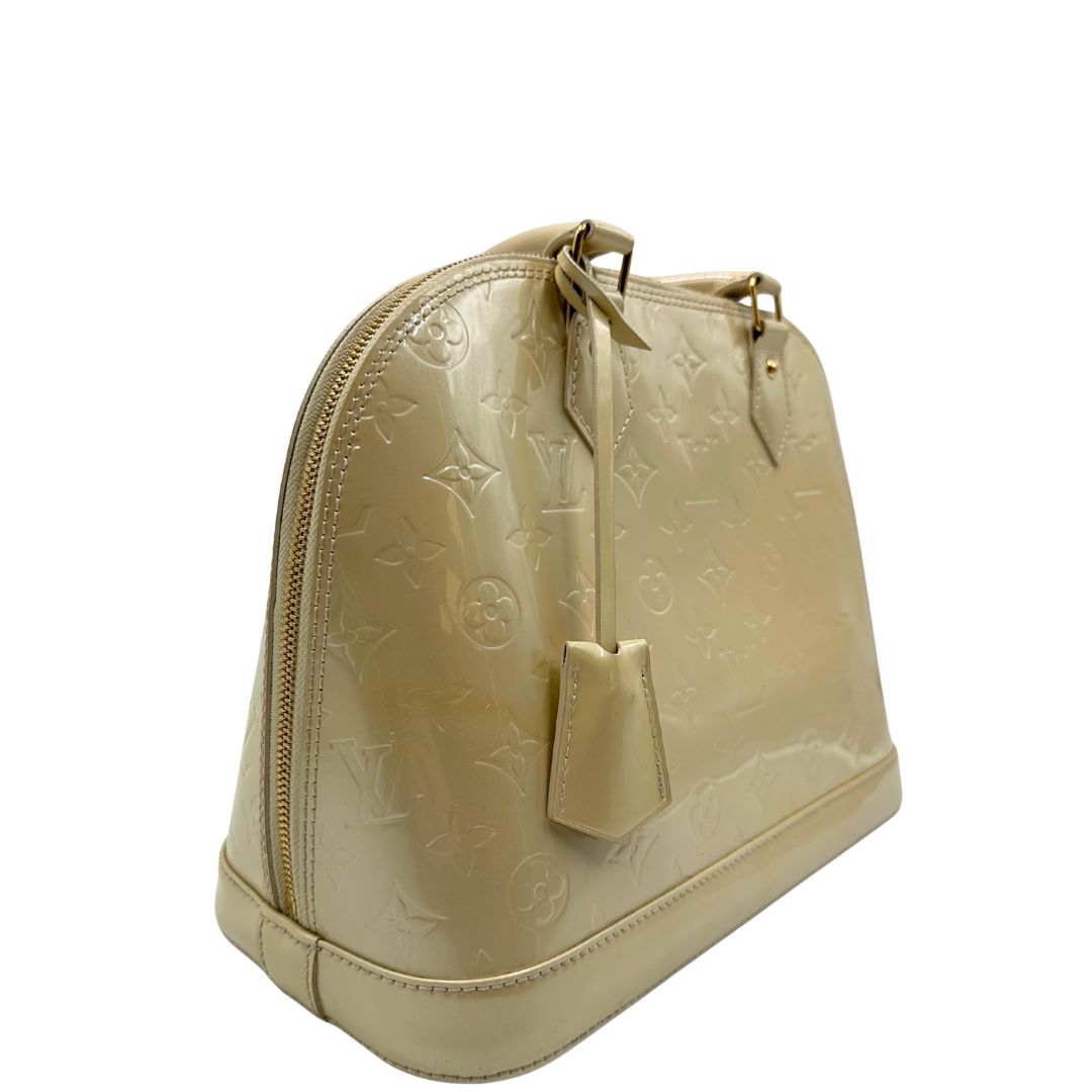 Laterale sinistro di una borsa Alma PM Louis Vuitton in pelle Vernis color noisette con parti metalliche dorate e doppi manici stondati; da portare a mano. Completa di lucchetto e chiavi. Indossabile a mano.