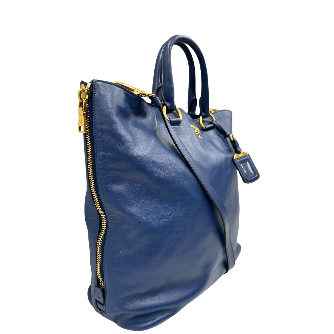 Laterale sinistro di una borsa Prada in pelle bluette con parti metalliche dorate; munita di un accessorio porta address, doppi manici stondati e una tracolla amovibile e regolabile. Completa di dustbag e card d'autenticità. Indossabile a mano oppure a tracolla.