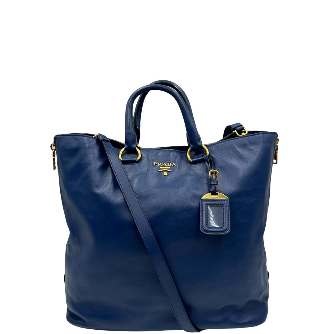 Front di una borsa Prada in pelle bluette con parti metalliche dorate; munita di un accessorio porta address, doppi manici stondati e una tracolla amovibile e regolabile. Completa di dustbag e card d'autenticità. Indossabile a mano oppure a tracolla.
