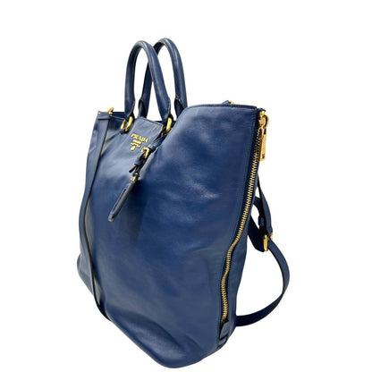 Laterale destro di una borsa Prada in pelle bluette con parti metalliche dorate; munita di un accessorio porta address, doppi manici stondati e una tracolla amovibile e regolabile. Completa di dustbag e card d'autenticità. Indossabile a mano oppure a tracolla.