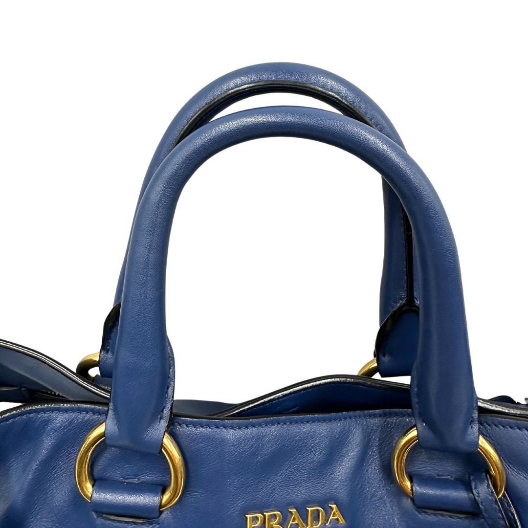 Manici di una borsa Prada in pelle bluette con parti metalliche dorate; munita di un accessorio porta address, doppi manici stondati e una tracolla amovibile e regolabile. Completa di dustbag e card d'autenticità. Indossabile a mano oppure a tracolla.