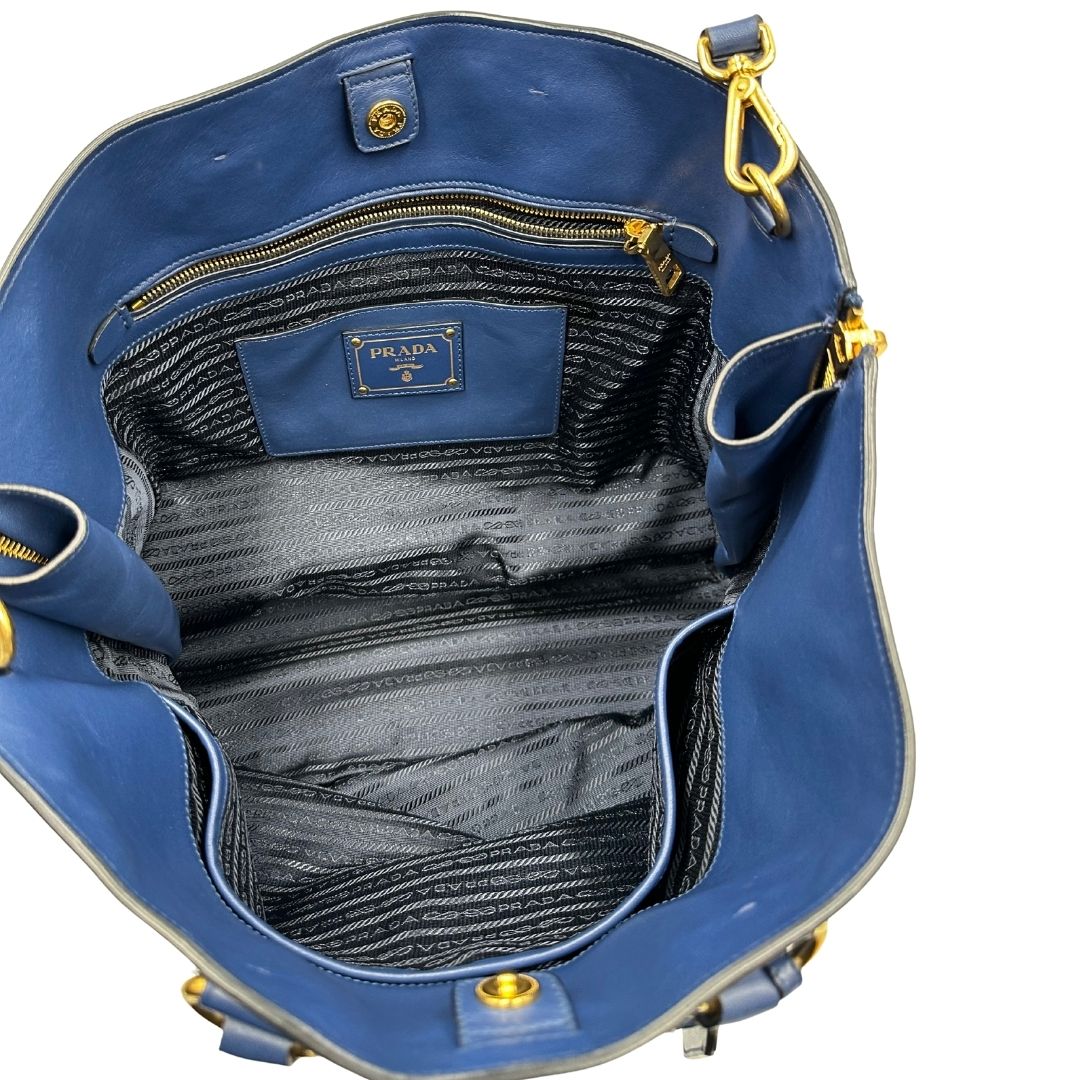 Interno di una borsa Prada in pelle bluette con parti metalliche dorate; munita di un accessorio porta address, doppi manici stondati e una tracolla amovibile e regolabile. Completa di dustbag e card d'autenticità. Indossabile a mano oppure a tracolla.