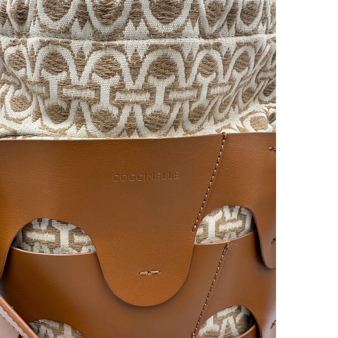 Logo di una borsa Coccinelle in pelle color cammello con parti metalliche argentate e interno in tessuto logato; munita di un manico singolo leggermente regolabile. Indossabile a spalla.