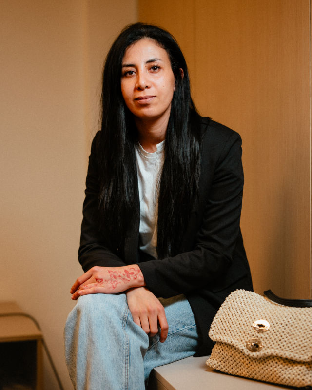 Donna seduta con giacca scura e jeans, tiene le mani sulle gambe con borsa chiara appoggiata accanto.