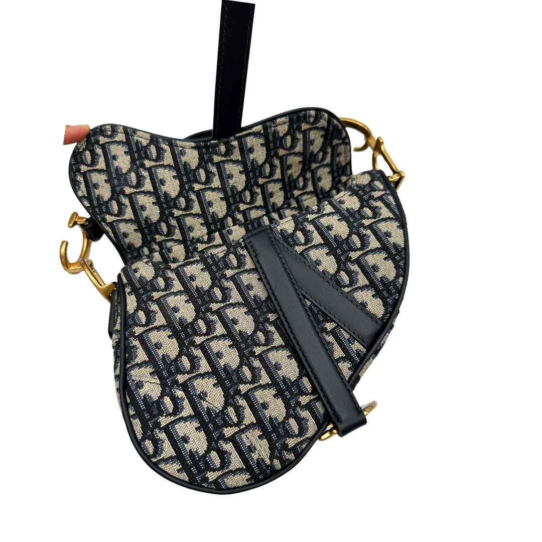 Borsa Saddle Dior in tessuto beige con trama Dior Oblique nera e blu, finiture in pelle blu notte e parti metalliche dorate. Munita di manico singolo e tracolla regolabile, e amovibile. Completa di corredo. Originale, usato, condizioni ottime. 