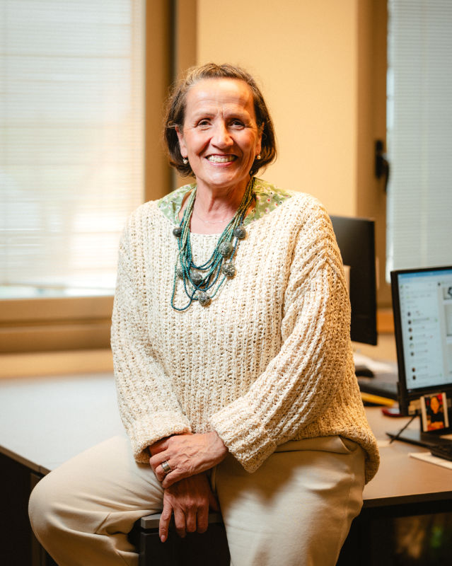 Donna seduta con un maglione chiaro e collana, sorridente in un ambiente d’ufficio luminoso.
