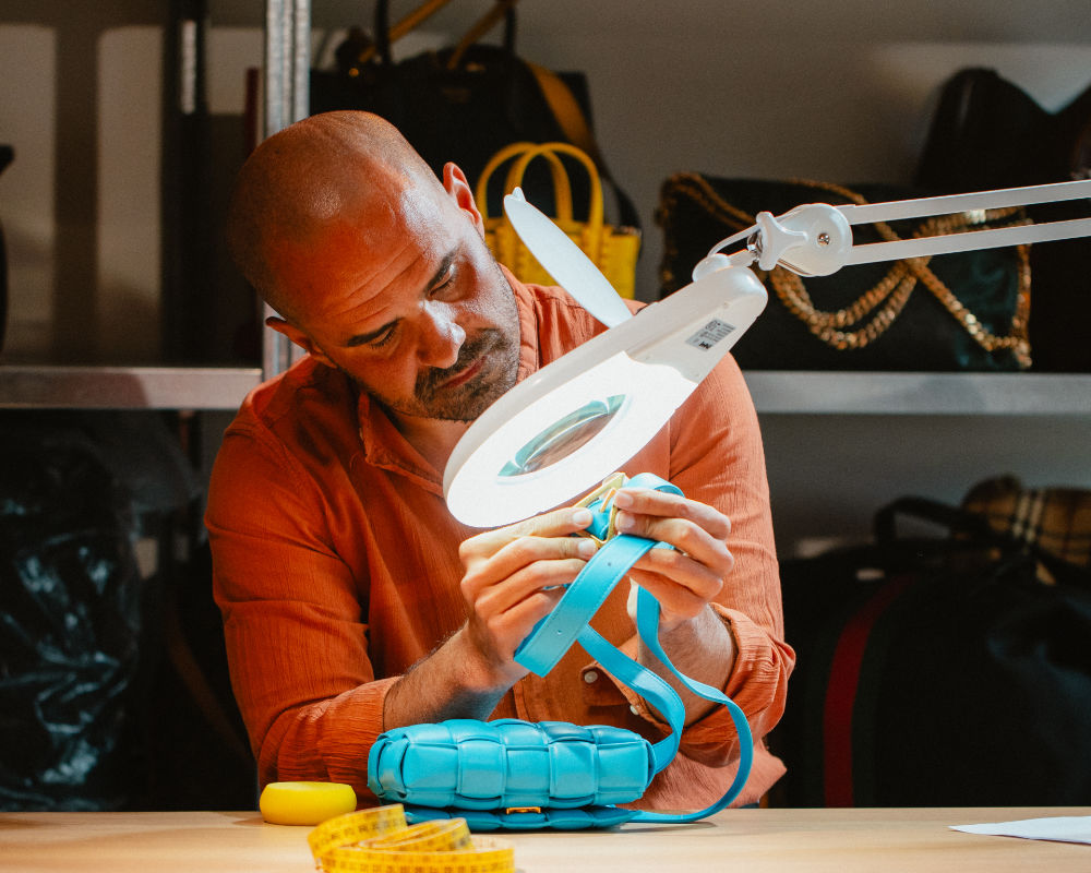 Uomo seduto a un banco da lavoro mentre osserva attentamente la fibbia di una borsa in pelle azzurra sotto una lampada con lente d’ingrandimento.