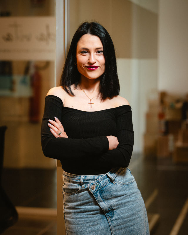 Donna in piedi con braccia incrociate, indossa un top nero e jeans, posa sorridente davanti all’obiettivo.