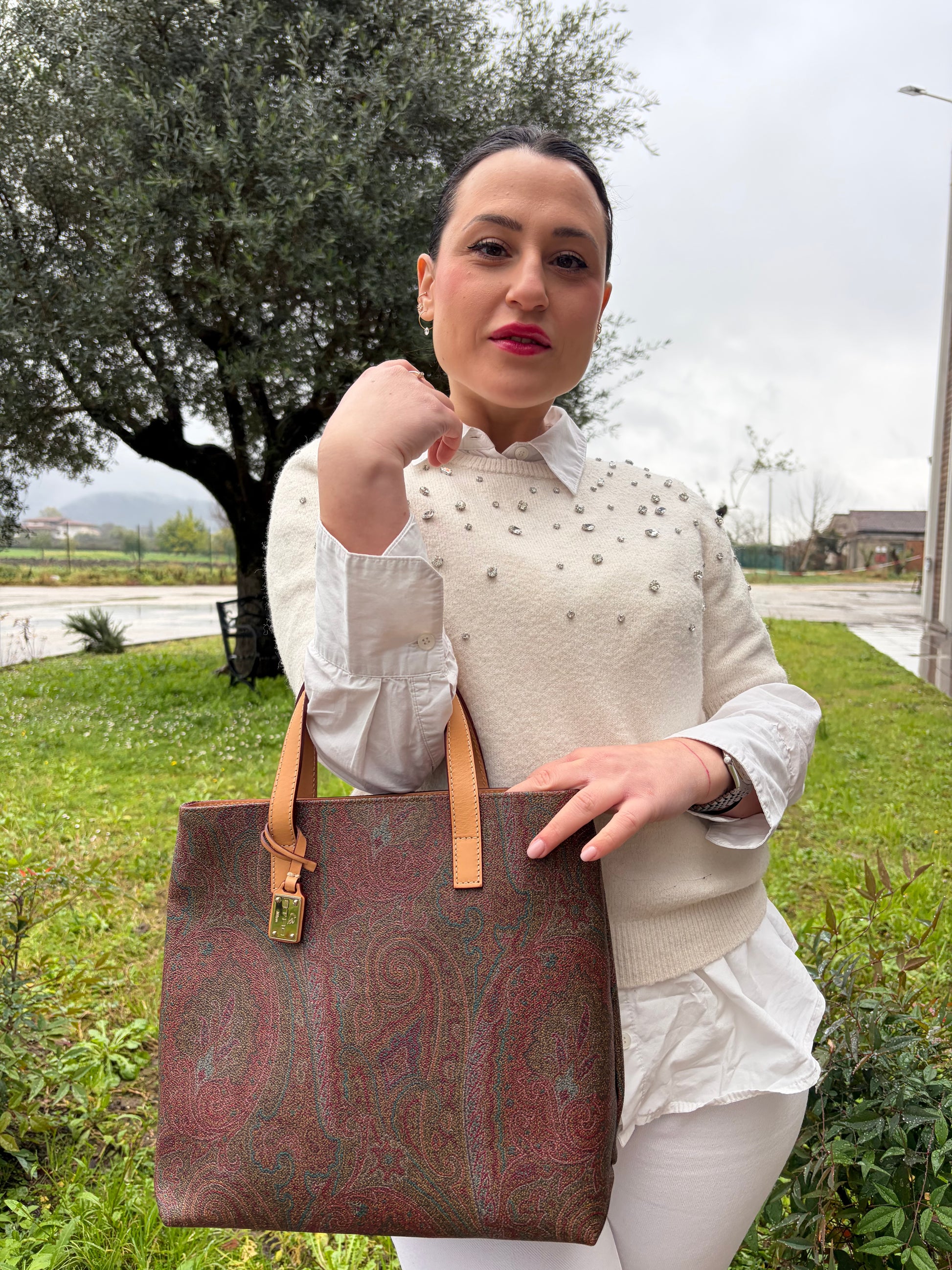 Ragazza che indossa una borsa Etro in canvas trama paisley in varie tonalità calde e parti in pelle beige, dettagli metallici dorati, munita di doppi manici. Indossabile a mano. Completa di dustbag e charm decorativo amovibile.