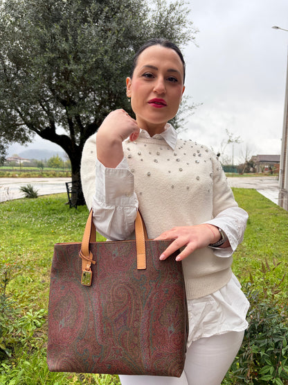 Ragazza che indossa una borsa Etro in canvas trama paisley in varie tonalità calde e parti in pelle beige, dettagli metallici dorati, munita di doppi manici. Indossabile a mano. Completa di dustbag e charm decorativo amovibile.