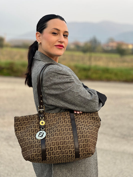 Ragazza che indossa una borsa Fendi in tessuto beige trama FF marrone, con finiture in pelle testa di moro. Dotata di doppi manici leggermente regolabili e parti metalliche dorate. Indossabile a spalla.