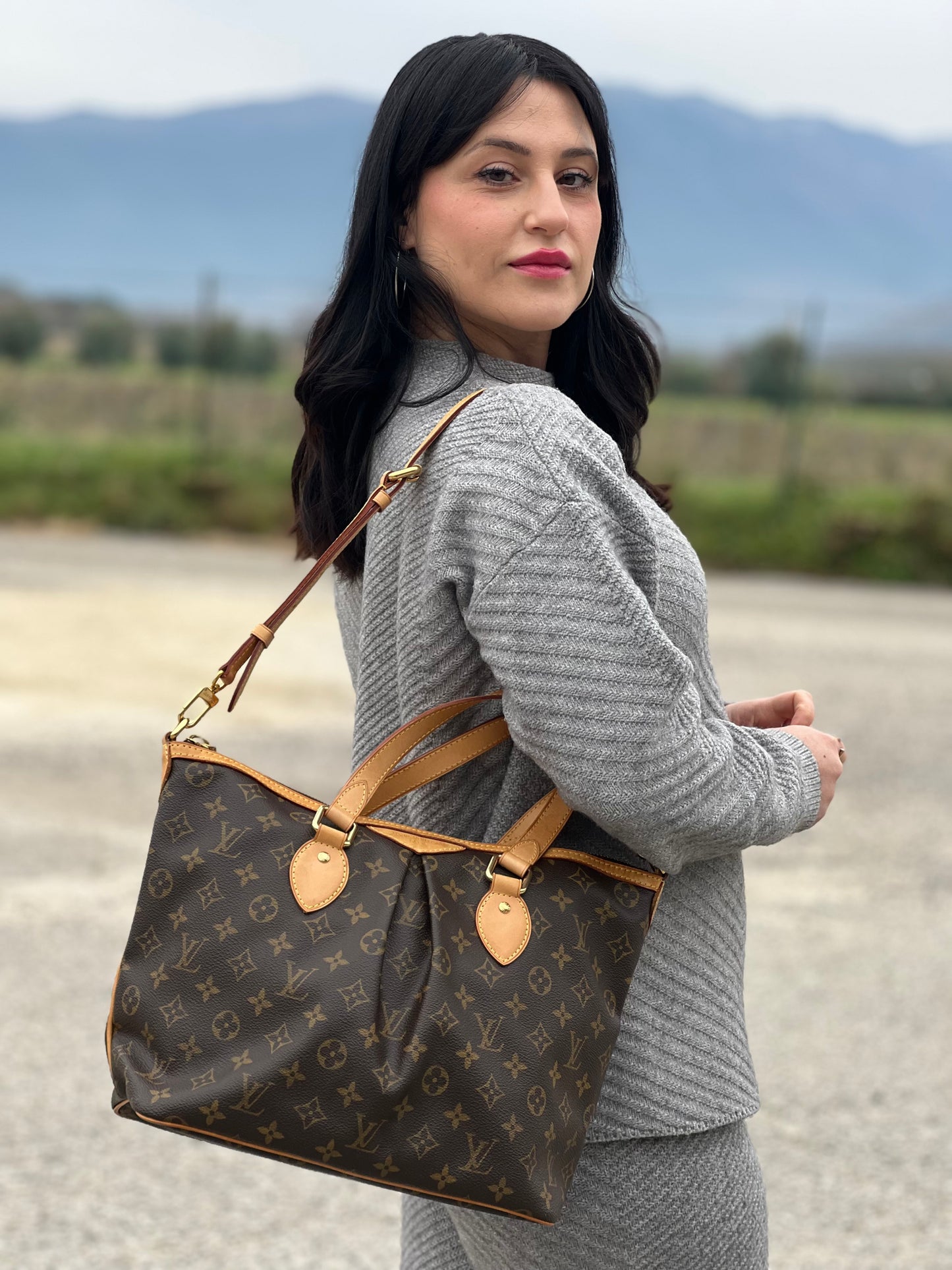 Ragazza che indossa una borsa Louis Vuitton in canvas marrone monogram con parti metalliche dorate e finiture in vacchetta naturale; munita di una tracolla amovibile e regolabile e doppi manici. Indossabile a spalla oppure a mano.
