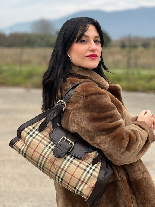 Ragazza che indossa una borsa Burberry in canvas beige, motivo Burberry check, con dettagli e finiture in pelle marroni testa di moro, parti metalliche dorate. Munita di doppi manici leggermente regolabili, indossabile a spalla. Completa di dustbag.