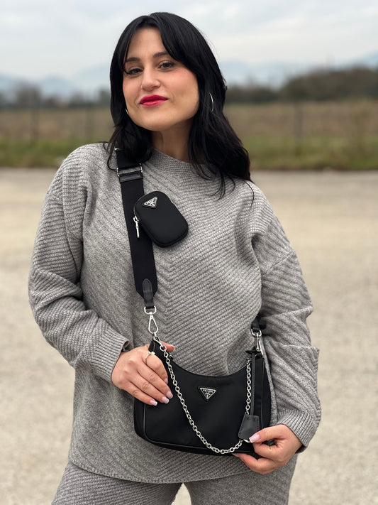 Ragazza che indossa una borsa Prada in nylon nero con parti metalliche argentate e finiture in pelle tono su tono; munita di una mini pouch amovibile, un accessorio portachiavi, manico in catena e una tracolla in tessuto amovibile e regolabile.