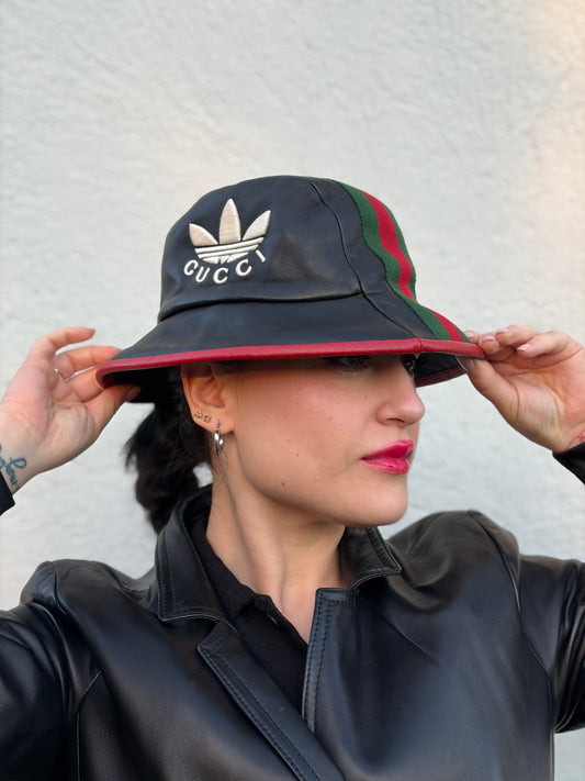 Ragazza che indossa un cappello Gucci x Adidas in pelle nera con logo frontale bianco avorio 'Gucci x Adidas'. Caratterizzato da fascia Web in tessuto verde/rosso e profili rossi.