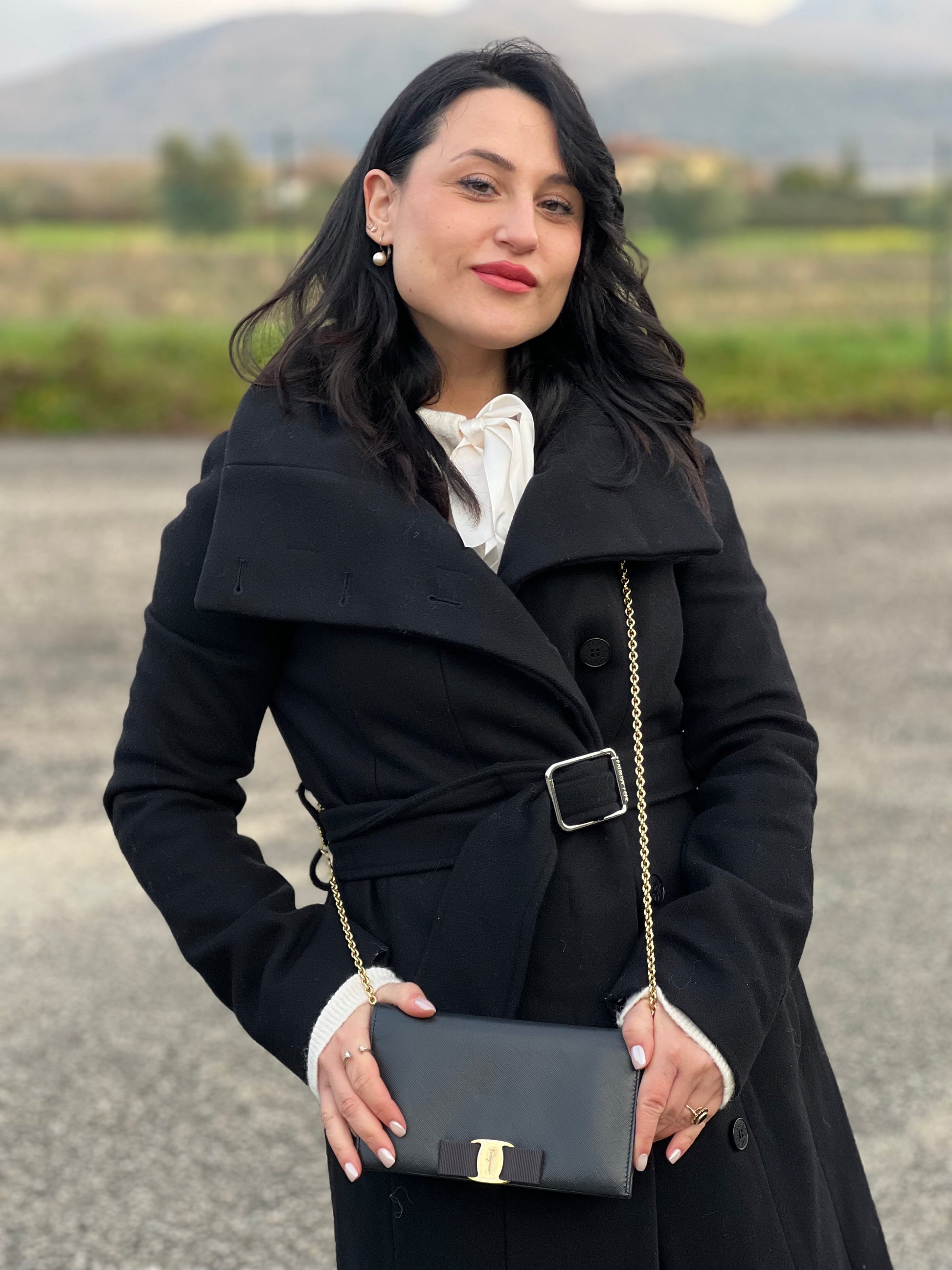 Ragazza che indossa un WOC Salvatore Ferragamo in pelle saffiano nera con parti metalliche oro. Munita di una tracolla in catena amovibile e non regolabile. Chiusura con fiocco. Completo di dustbag. Indossabile a tracolla.