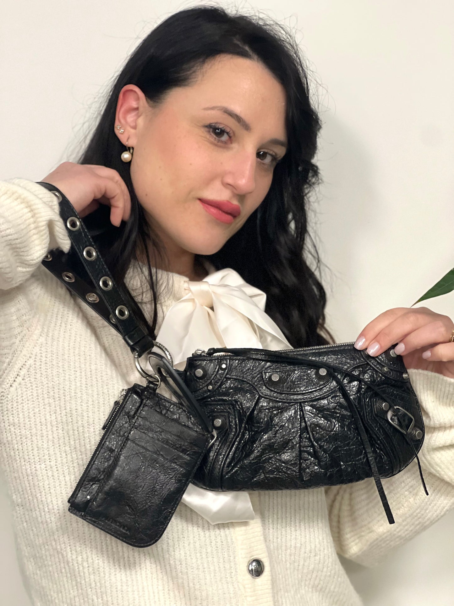 Ragazza che indossa una pochette Balenciaga in pelle effetto invecchiato nera, con parti metalliche argentate; munita di uno specchietto a forma di cuore, polsino amovibile e un mini porta monete e carte. Completa di dustbag.