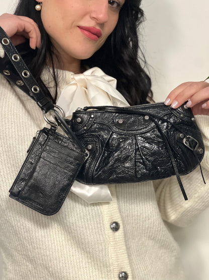 Ragazza che indossa una pochette Balenciaga in pelle effetto invecchiato nera, con parti metalliche argentate; munita di uno specchietto a forma di cuore, polsino amovibile e un mini porta monete e carte. Completa di dustbag.