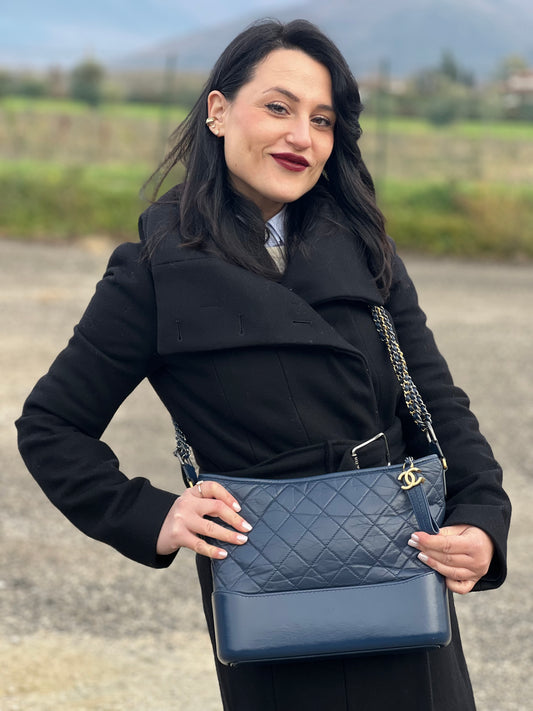Ragazza che indossa una borsa Chanel Gabrielle in pelle matelassé blu navy con parti metalliche dorate e argentate. Munita di una tracolla in catena e pelle intrecciata con spallaccio. Tirante delle cerniera impreziosito dall'iconico logo CC in metallo dorato, completa di corredo.