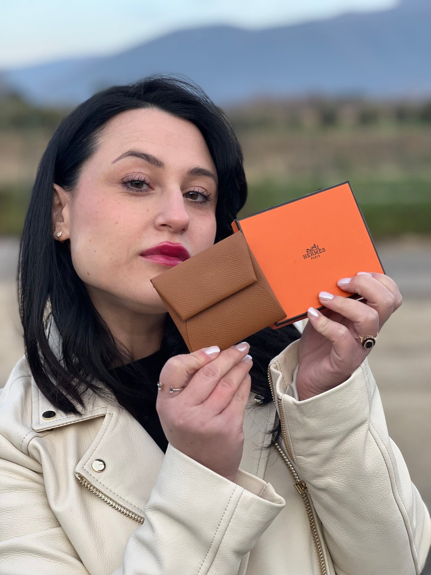 Ragazza che mostra un mini portamonete Hermès in pelle color cammello, con chiusura a bottone. Completo di scatola.