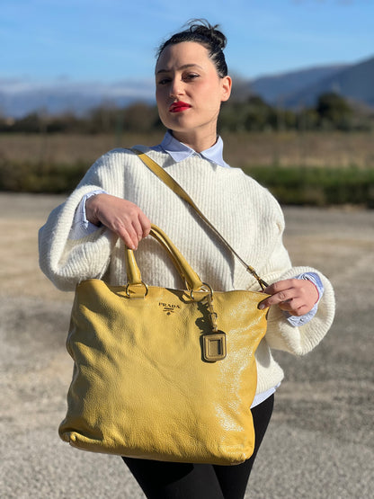 Ragazza che indossa una Borsa Prada in pelle lucida color mustard con parti metalliche dorate; munita di un accessorio porta address e doppi manici stondati. Indossabile a mano oppure a tracolla. Originale, usata, di lusso, in ottime condizioni