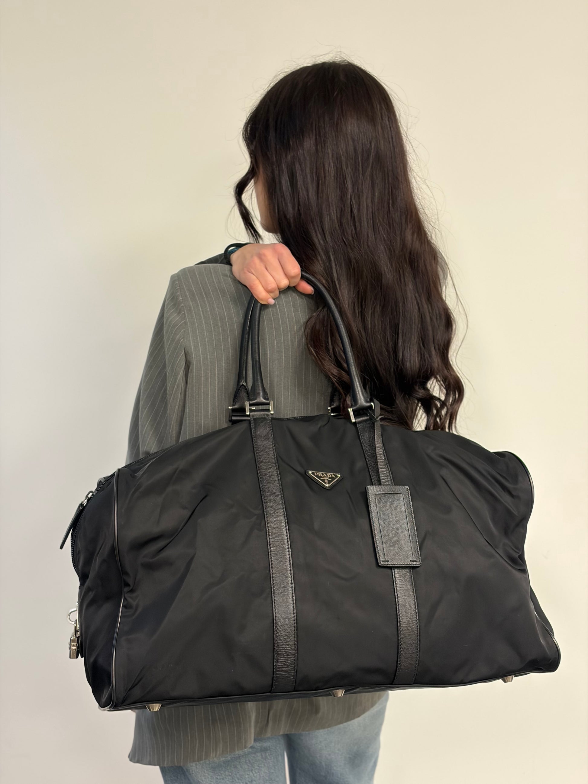 Ragazza che mostra un borsone Prada in nylon nero con finiture in pelle Saffiano tono su tono e parti metalliche argentate. Completo di dustbag, tag porta address e lucchetto. Indossabile a mano.