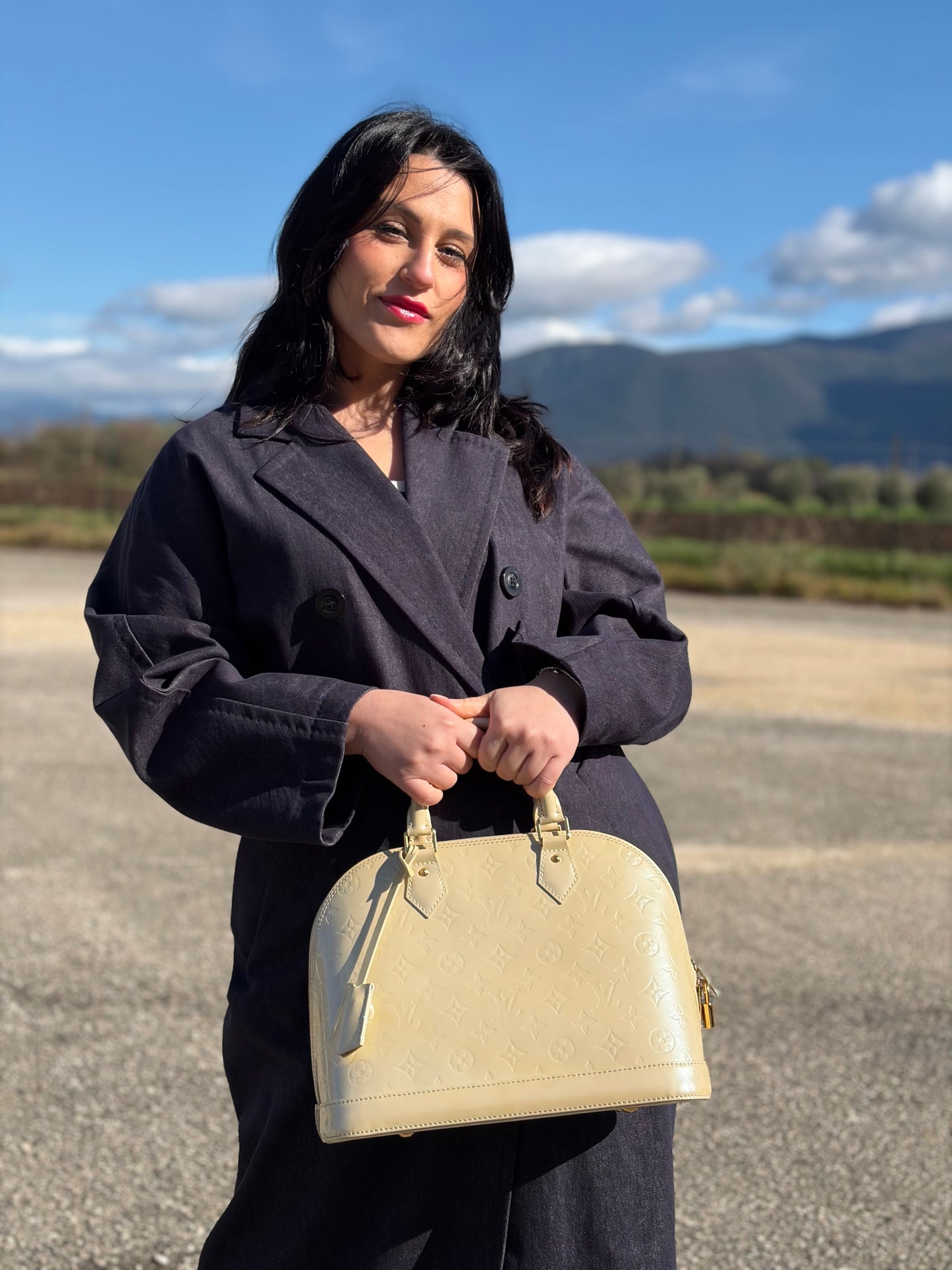 Ragazza che indossa una borsa Alma PM Louis Vuitton in pelle Vernis color noisette con parti metalliche dorate e doppi manici stondati; da portare a mano. Completa di lucchetto e chiavi. Indossabile a mano.