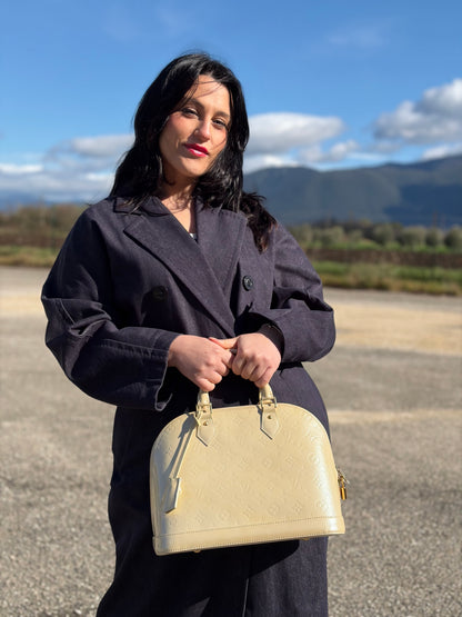 Ragazza che indossa una borsa Alma PM Louis Vuitton in pelle Vernis color noisette con parti metalliche dorate e doppi manici stondati; da portare a mano. Completa di lucchetto e chiavi. Indossabile a mano.
