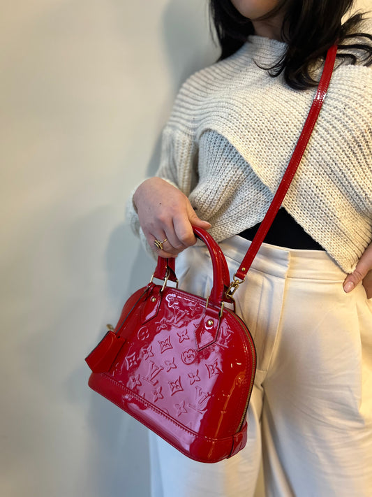 Ragazza che indossa una borsa Louis Vuitton Alma BB in pelle vernis rossa con trama monogram empreinte e parti metalliche dorate. Munita di doppi manici stondati e tracolla amovibile, dotata di clochette porta chiavi. Completa di scatola, dustbag, lucchetto e chiavi.