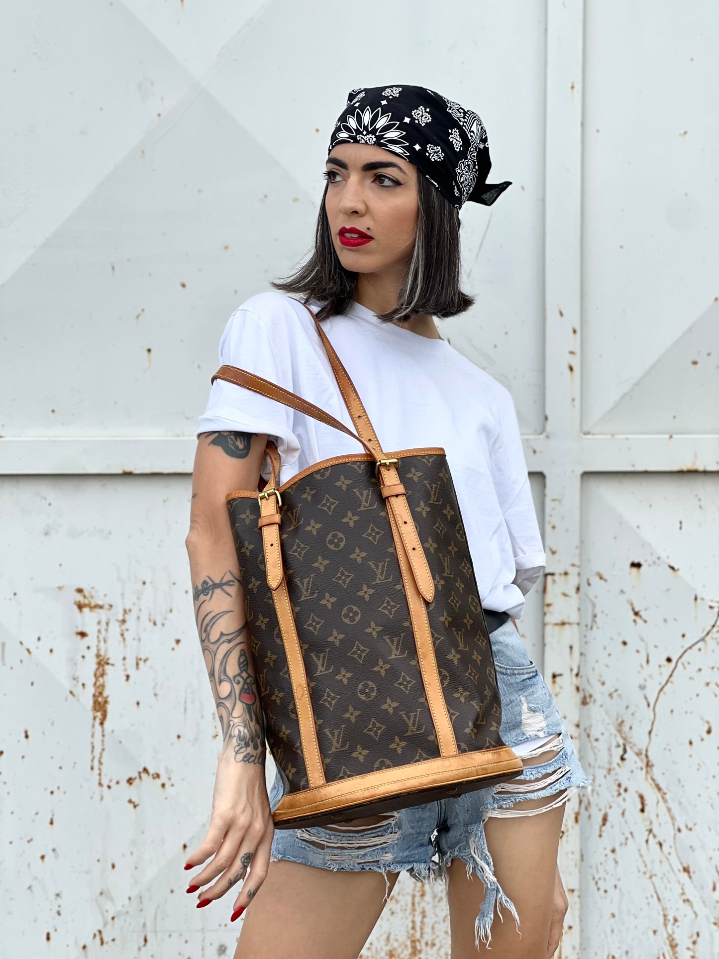 Ragazza che indossa un bucket Louis Vuitton in canvas marrone monogram con finiture in vacchetta naturale, manici regolabili con fibbie e parti metalliche dorate.