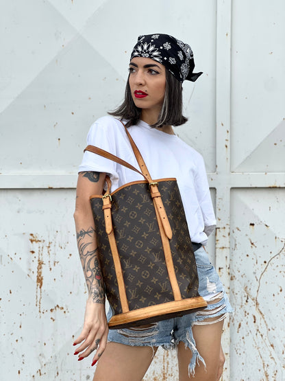 Ragazza che indossa un bucket Louis Vuitton in canvas marrone monogram con finiture in vacchetta naturale, manici regolabili con fibbie e parti metalliche dorate.