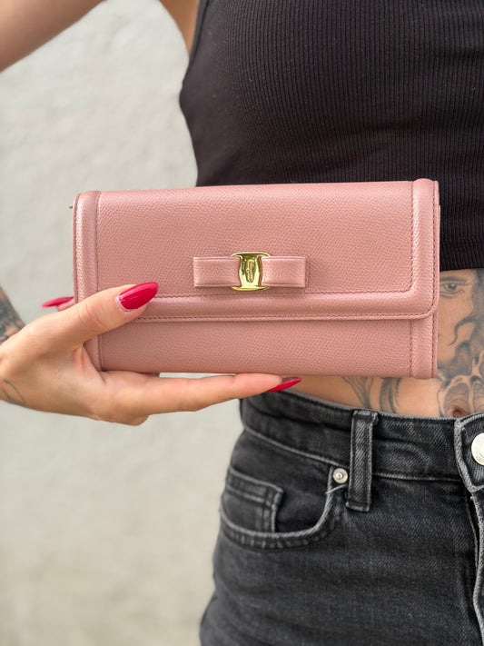 Ragazza che mostra un portafoglio Ferragamo in pelle rosa, dotata di un porta tessera estraibile. Iconico fiocco del brand sul front, usato, originale, second hand, in buone condizioni.