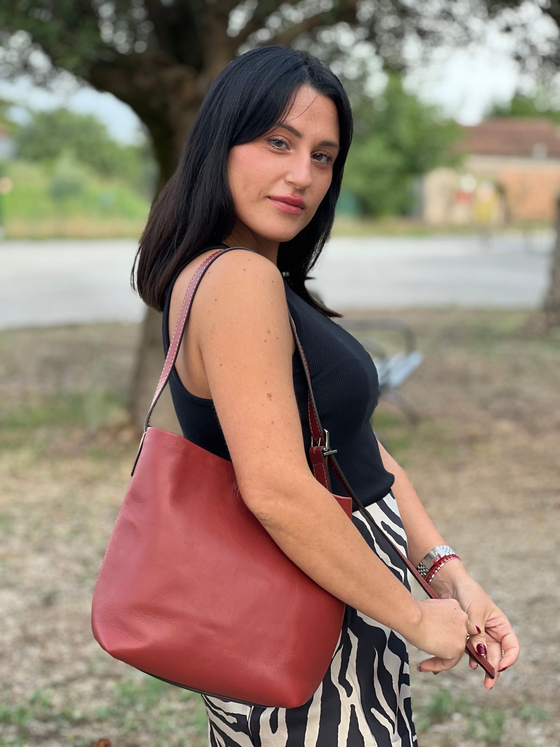 Ragazza che indossa una borsa a secchiello Hags in pelle color marrone bruciato con manico regolabile. Parti metalliche argentate. Indossabile a spalla o a tracolla. Completa di dustbag.