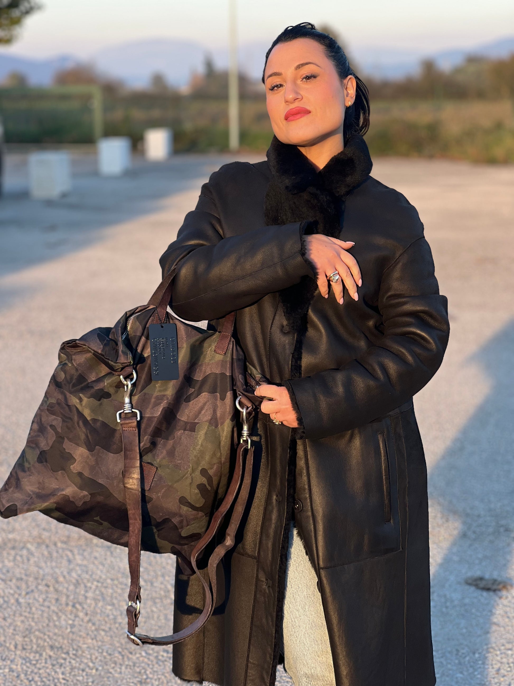 Ragazza che indossa una tote Campomaggi in nylon trama camouflage, con finiture in pelle testa di moro e parti metalliche argentate. Munita di tracolla amovibile e regolabile. Completa di dustbag.