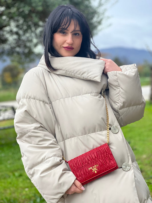 Ragazza che indossa un wallet Prada in pelle goffrata rossa, impreziosito da logo lettering sul front e specchietto interno; munito di una tracolla in catena amovibile e non regolabile. Completo di scatola.