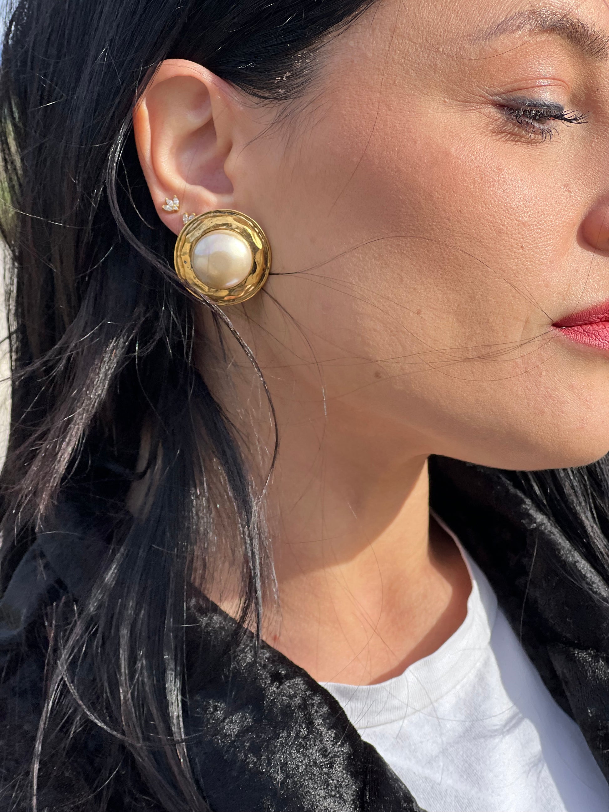 Ragazza che indossa degli orecchini Chanel vintage a clip, caratterizzati da una montatura in metallo dorato martellata che incornicia una grande perla artificiale centrale dal finish iridescente. Completi di scatola.