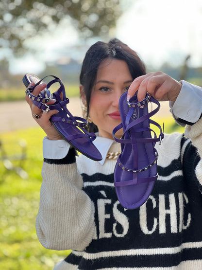 Ragazza che mostra dei sandali Tod’s in pelle viola con fasce incrociate e borchie metalliche argentate. Presentano una chiusura con cinturino e la suola con i classici gommini in gomma. Completi di scatola e dustbag.