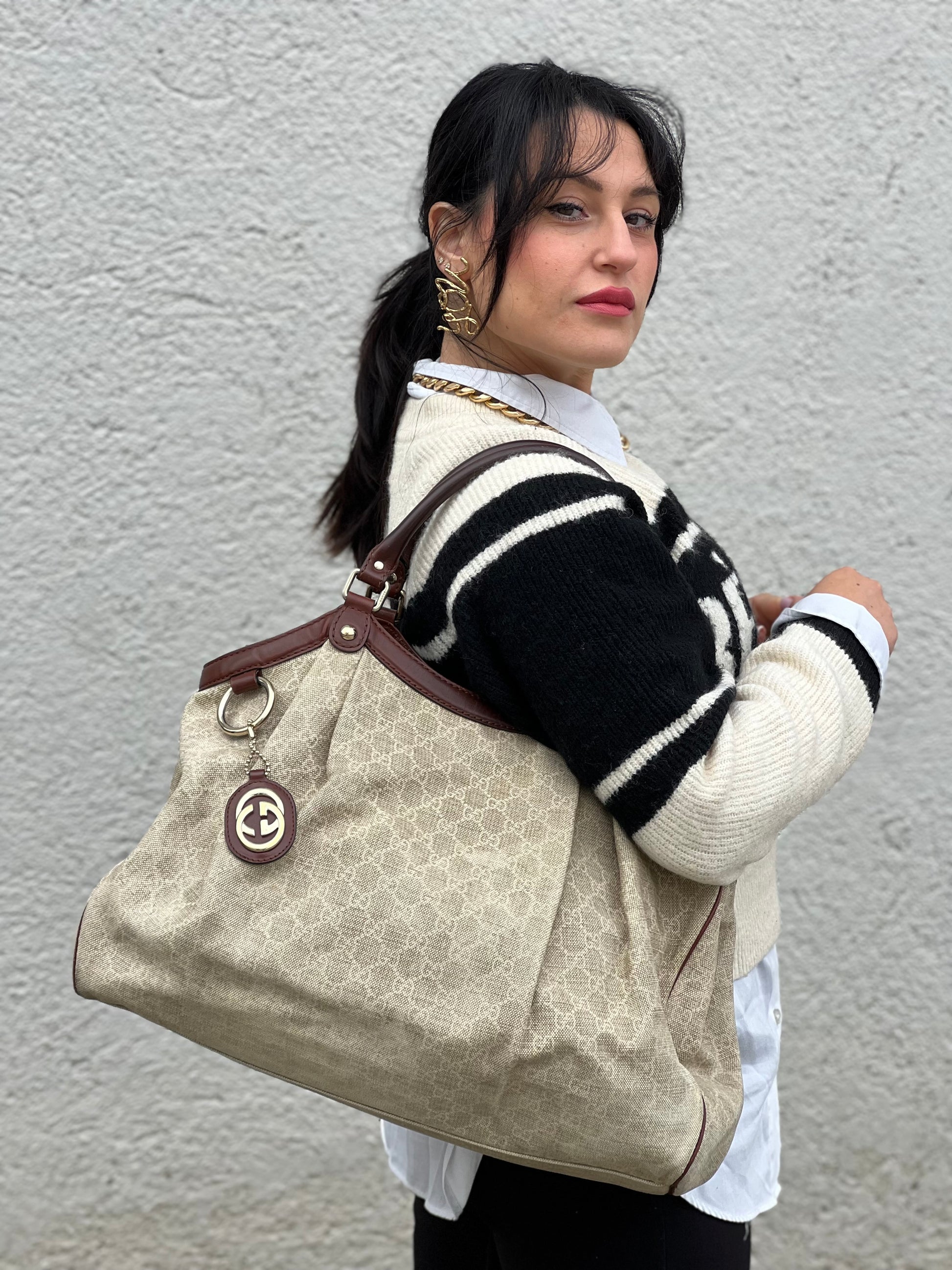 Ragazza che indossa una borsa Gucci Sukey in tessuto beige GG con finiture in pelle marrone mogano, doppi manici stondati e ciondolo con doppie G incrociate. Rifinita con parti metalliche dorate. Indossabile a spalla.