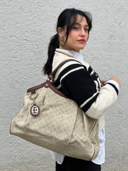 Ragazza che indossa una borsa Gucci Sukey in tessuto beige GG con finiture in pelle marrone mogano, doppi manici stondati e ciondolo con doppie G incrociate. Rifinita con parti metalliche dorate. Indossabile a spalla.