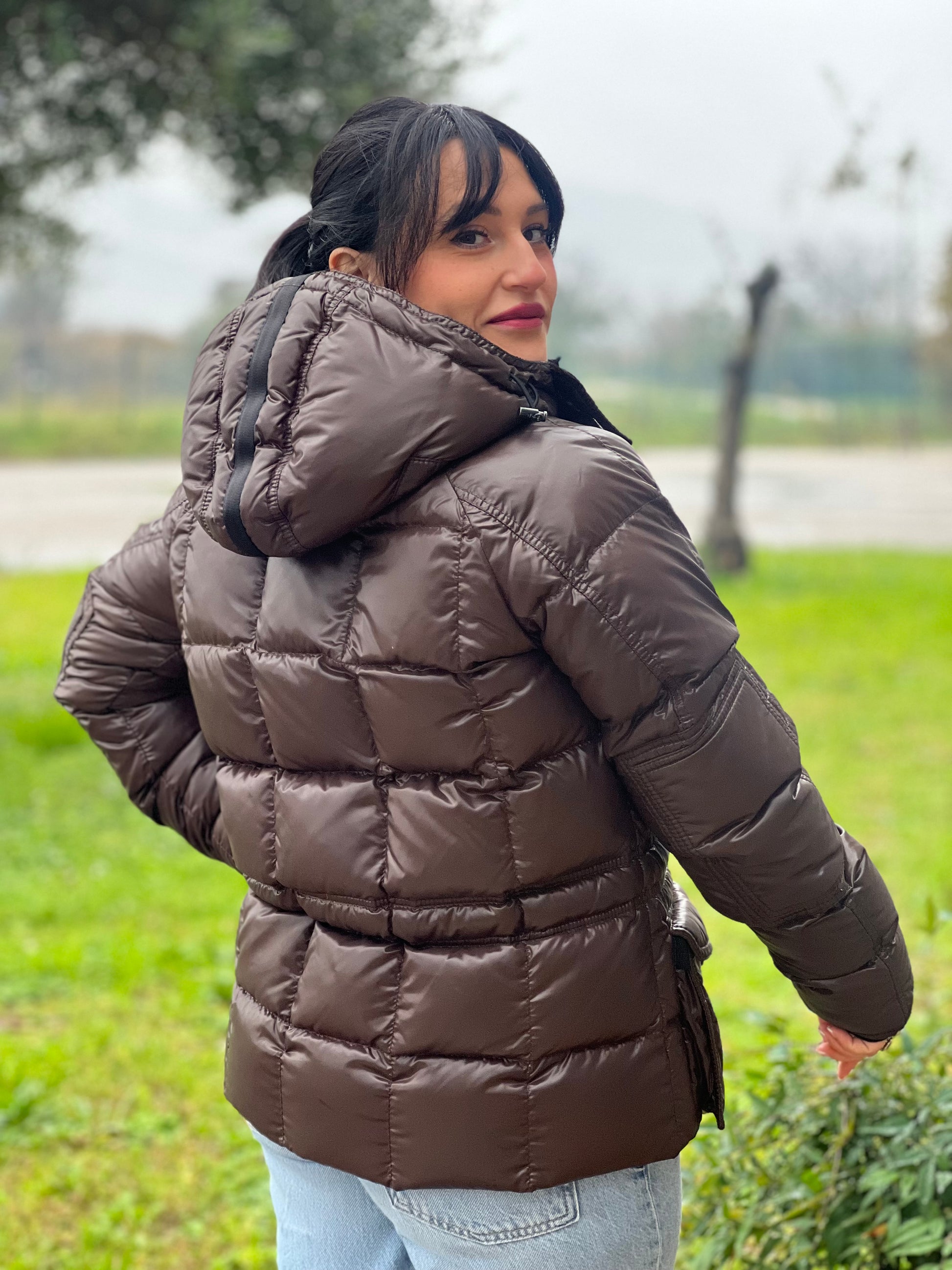 Ragazza che indossa un piumino Moncler in tessuto lucido color marrone, con parti metalliche effetto renio, in 100% poliammide, caratterizzato da imbottitura trapuntata, chiusura con zip e bottoni logati, cappuccio regolabile e polsini interni a coste per una maggiore protezione dal freddo.