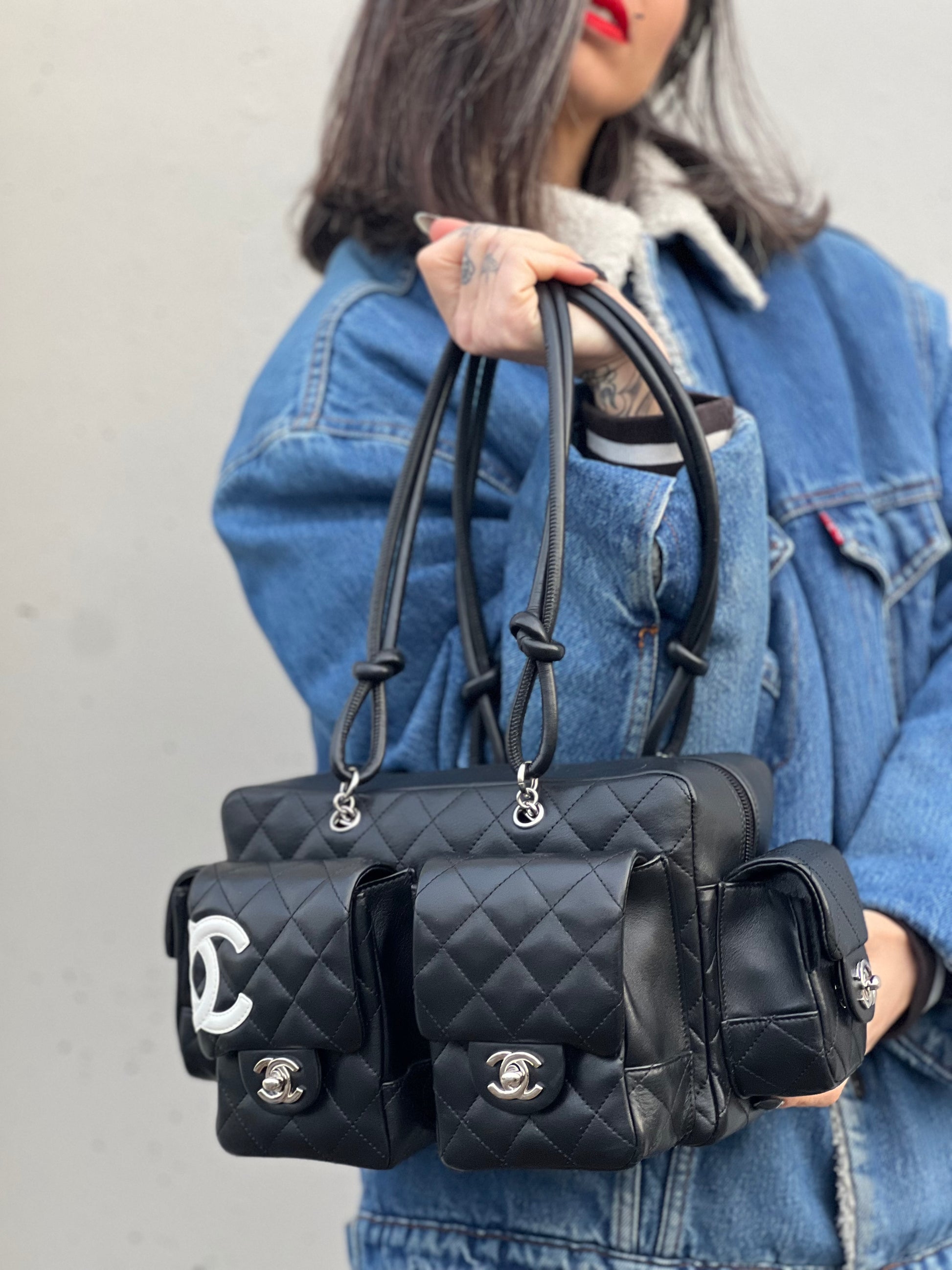 Ragazza che indossa una borsa Cambon Reporter Chanel in pelle trapuntata nera e doppie C bianche sul front, con tasche per accessori e doppi manici medio lunghi, rifinita con parti metalliche argentate. Completa di dustbag e card d'autenticità. Indossabile a spalla.