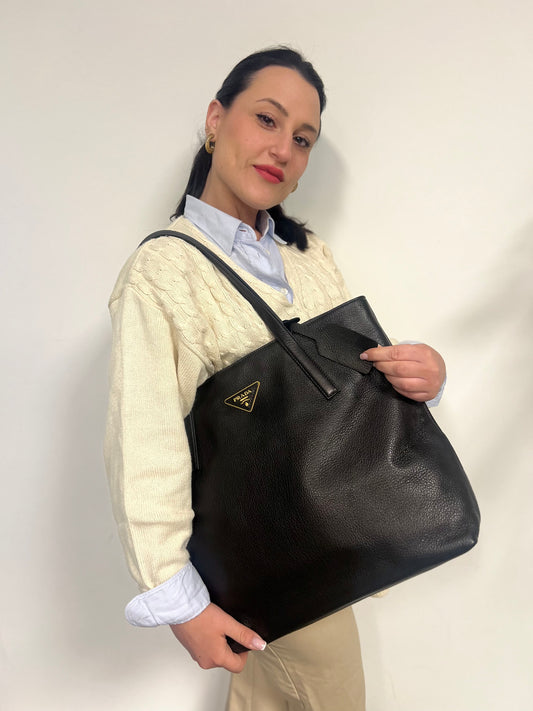 Ragazza che indossa una borsa tote Prada in pelle martellata nera con parti metalliche dorate; munita di doppi manici. Indossabile a spalla.