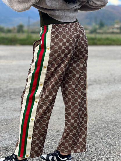 Ragazza che indossa un pantalone tuta Gucci elasticizzato modello Interlocking G in 55% poliestere e 45% cotone, marrone e avorio con motivo GG all-over e bande laterali con bottoni automatici che si aprono fino ai fianchi, con strisce Web verde/rosse.