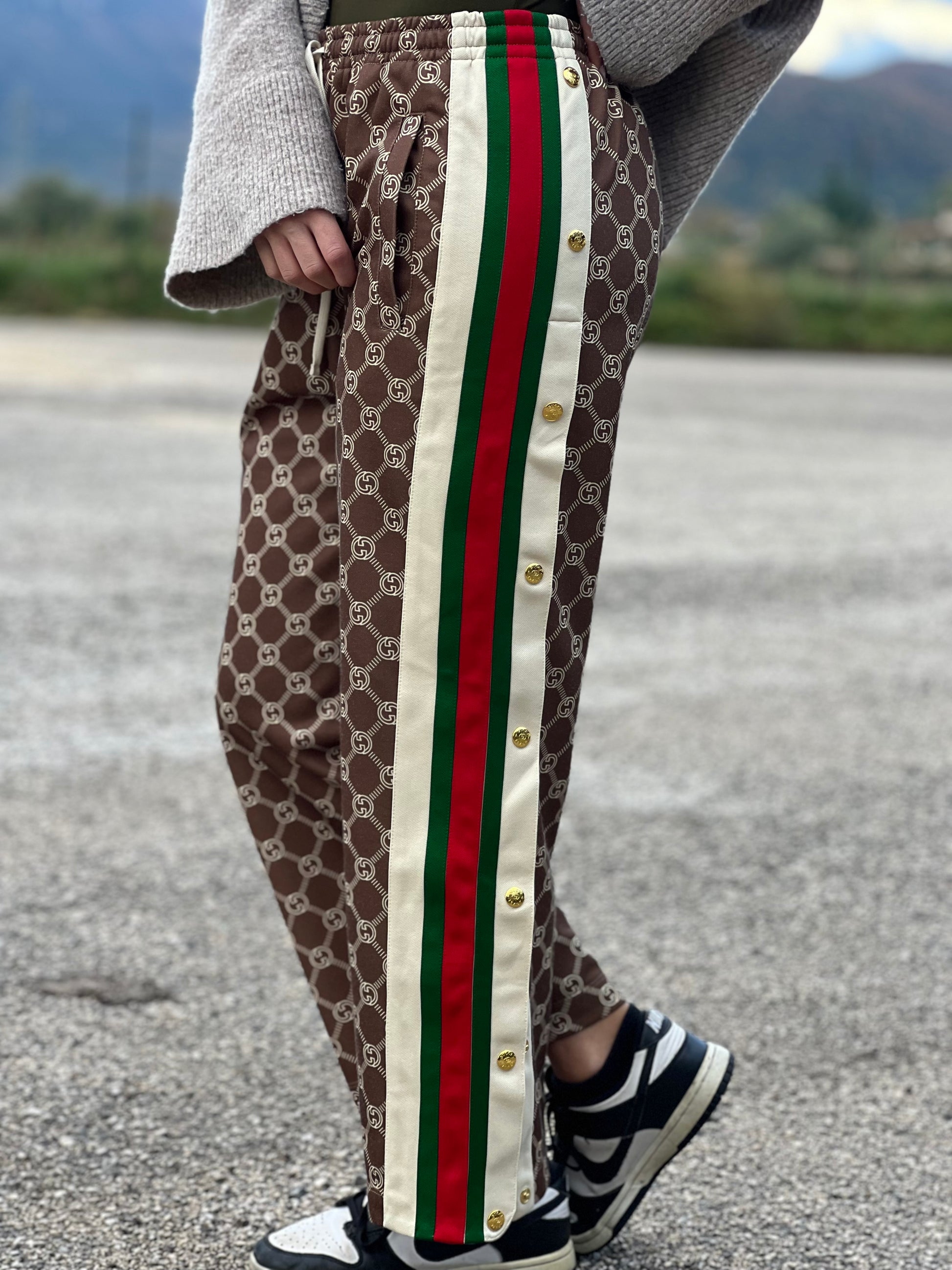 Ragazza che indossa un pantalone tuta Gucci elasticizzato modello Interlocking G in 55% poliestere e 45% cotone, marrone e avorio con motivo GG all-over e bande laterali con bottoni automatici che si aprono fino ai fianchi, con strisce Web verde/rosse.
