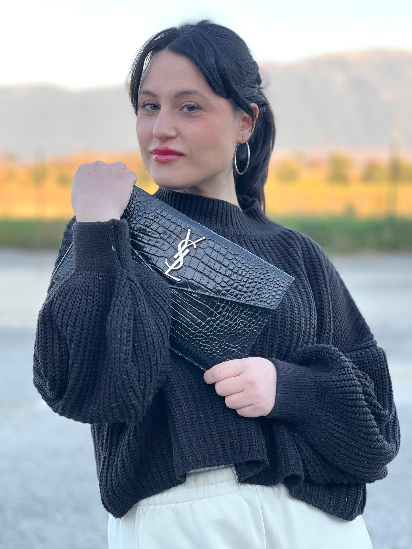 Ragazza che indossa una pochette Saint Laurent in pelle nera lucida effetto cocco, impreziosita dal logo Cassandre sul front in metallo argentato. Munita di dustbag.