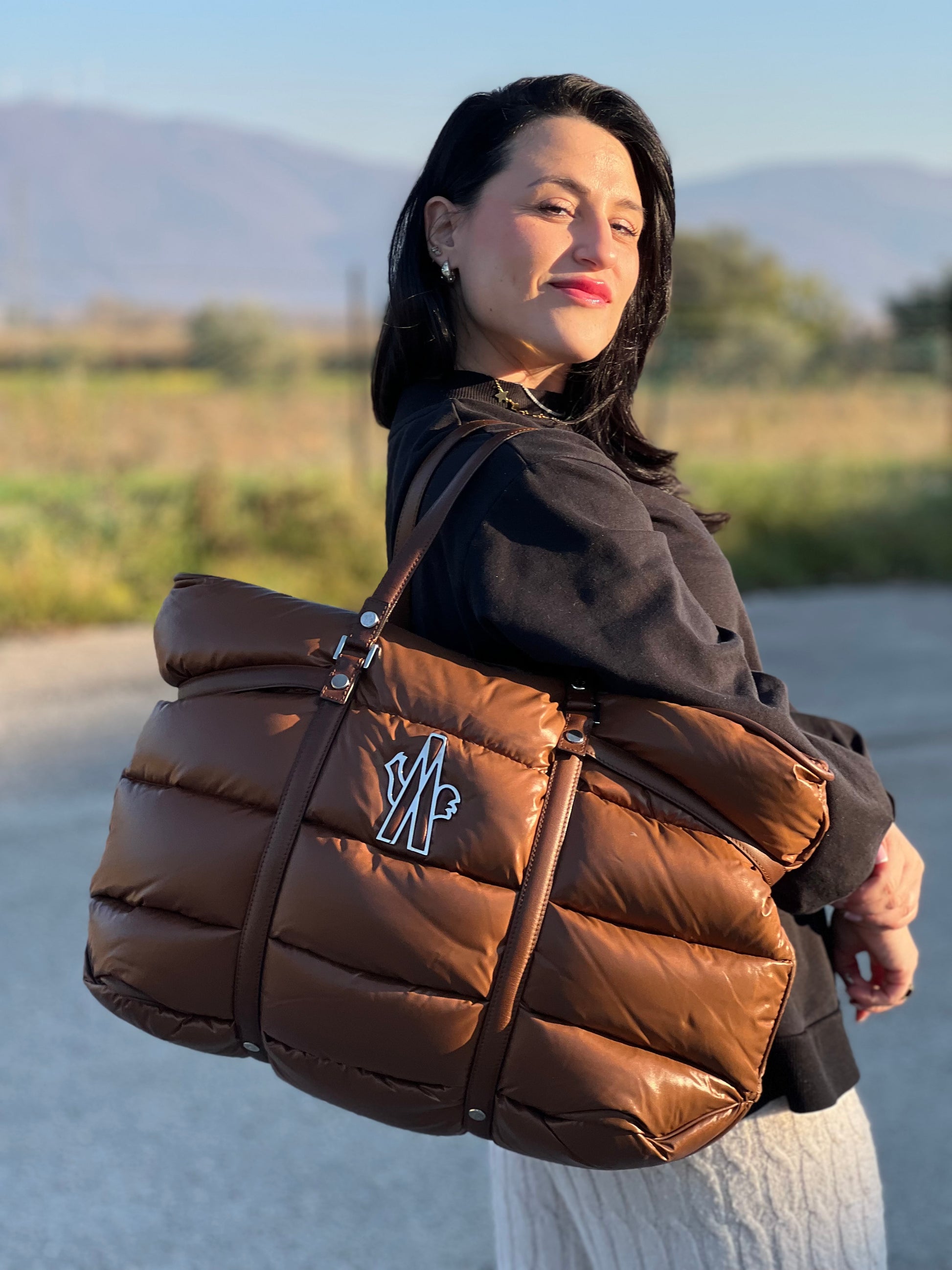 Ragazza che indossa una borsa Moncler in poliuretano color caramello effetto imbottito e finiture in pelle tono su tono, con metalleria dorata. Munita di cartellino. Da indossare a spalla.